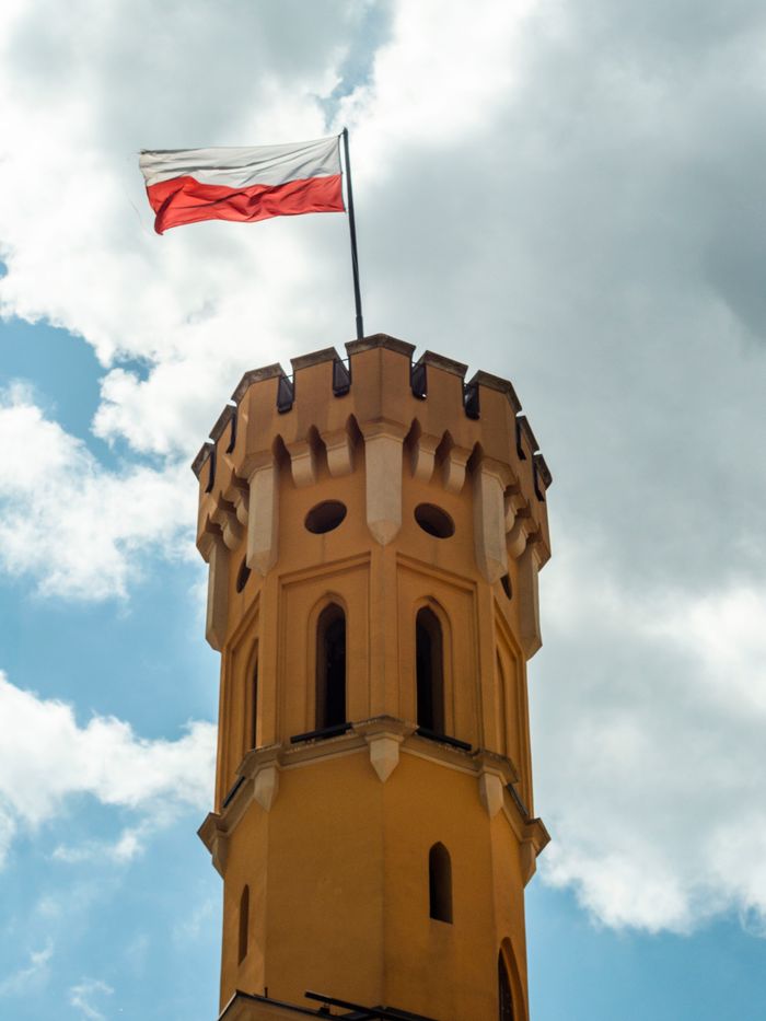 Historic tower with Polish flag waving against a bright blue sky and clouds.